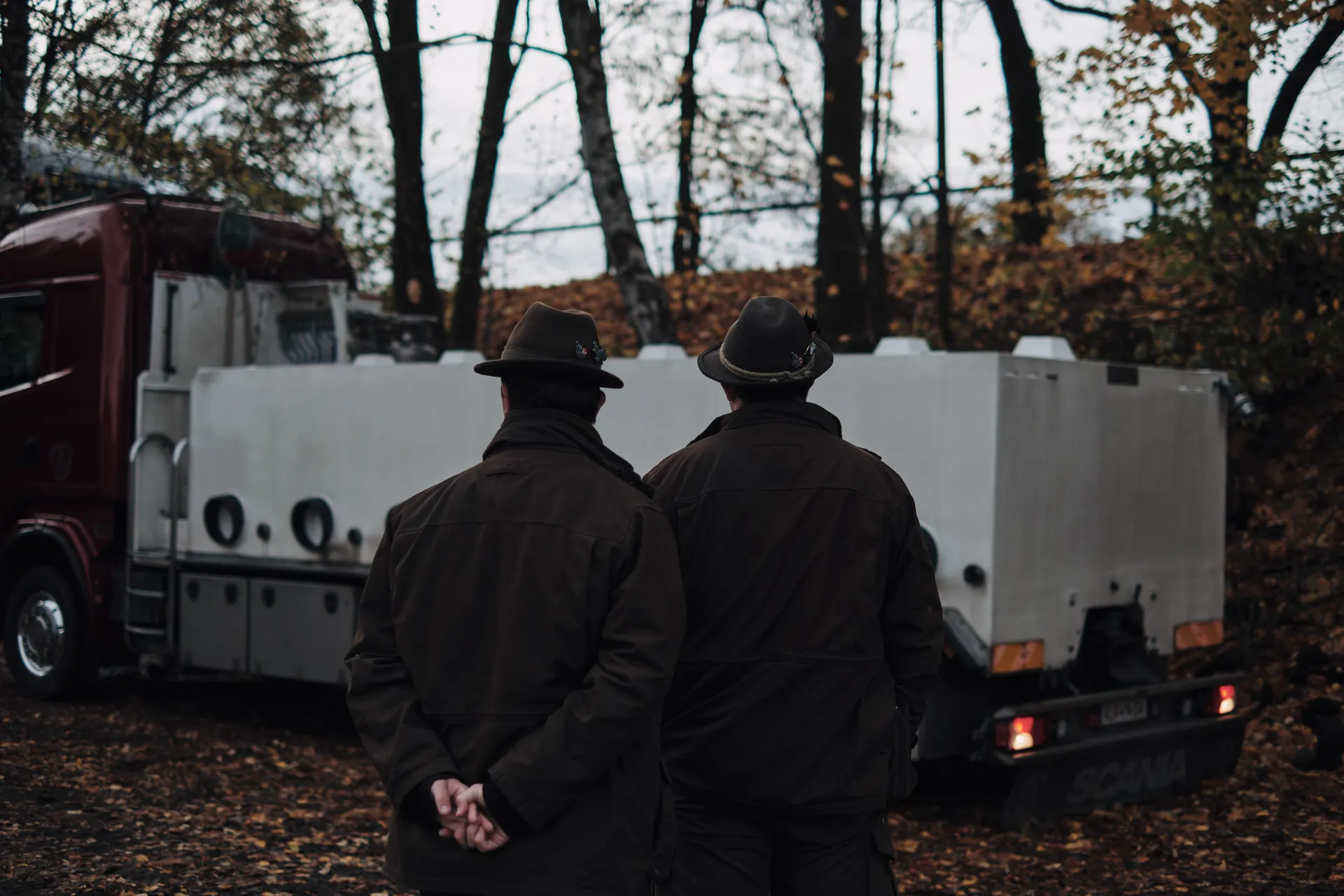 Zwei Männer mit Lodenjanker und Filzhut beobachten den Fischtransporter am Ufer.